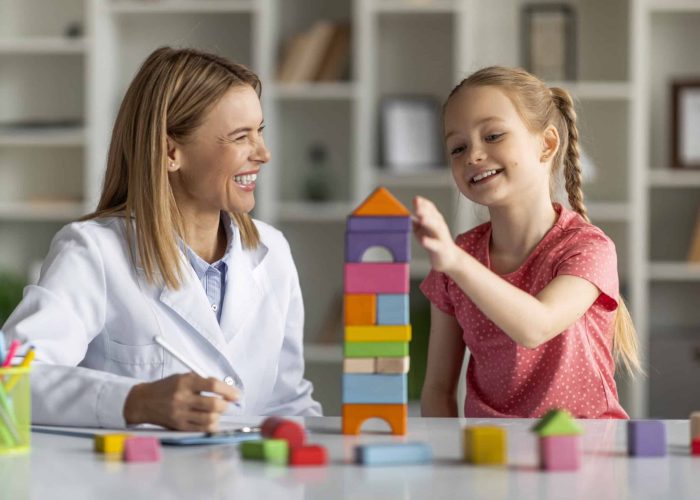 Child Development Specialist Looking At Little Girl Playing With Colorful Wooden Bricks During Session In Office, Professional Therapist Lady Having Lesson With Adorable Smiling Female Child